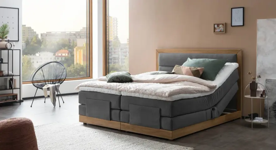 Modernes Boxspringbett mit Holzrahmen und grauem Stoffbezug. Die Matratze wirkt etwas zu hart, aber die Kissen laden zum Entspannen ein.