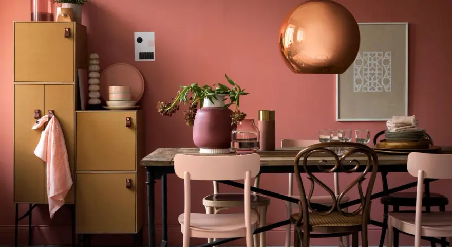 Ein Esszimmer mit altrosa Wänden. Ein kupferfarbener Lampenschirm und ein Holzmöbelstück mit gelben Türen. Welche Farbe zu altrosa? Zarte Rosatöne und Erdtöne passen gut.