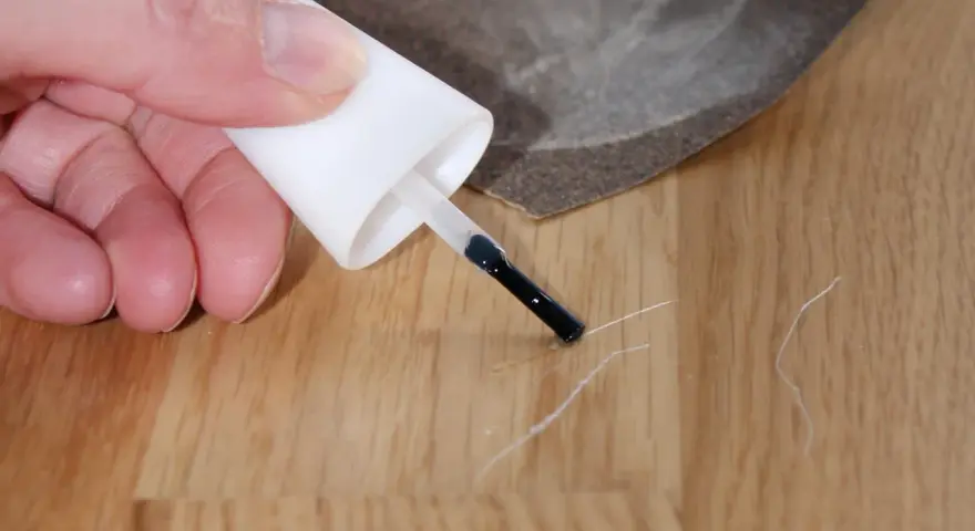 Kleine Kratzer im Parkett reparieren: Ein Finger hält einen Lackstift über eine Holzoberfläche, daneben liegt Schleifpapier.