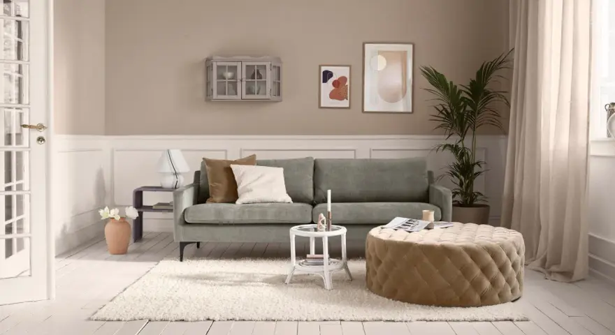 Ein Wohnzimmer mit salbeigrünem Sofa. Braune und cremefarbene Kissen, ein beiger Hocker und eine helle Wand zeigen, welche Farbe passt zu Salbei.