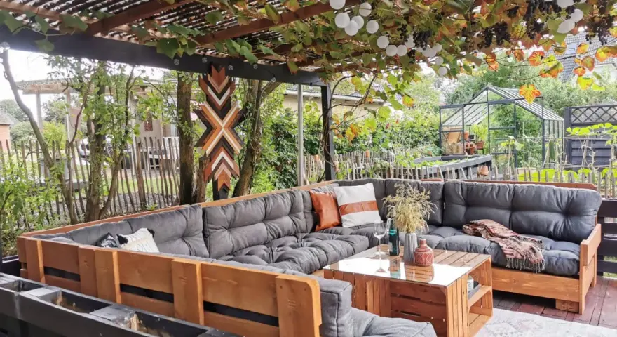 Gemütliche Paletten Lounge Ideen für den Garten. Ein großer Ecksofa aus Paletten mit grauen Polstern und Holztisch. Weinreben und Lichterketten überdachen den Sitzbereich.