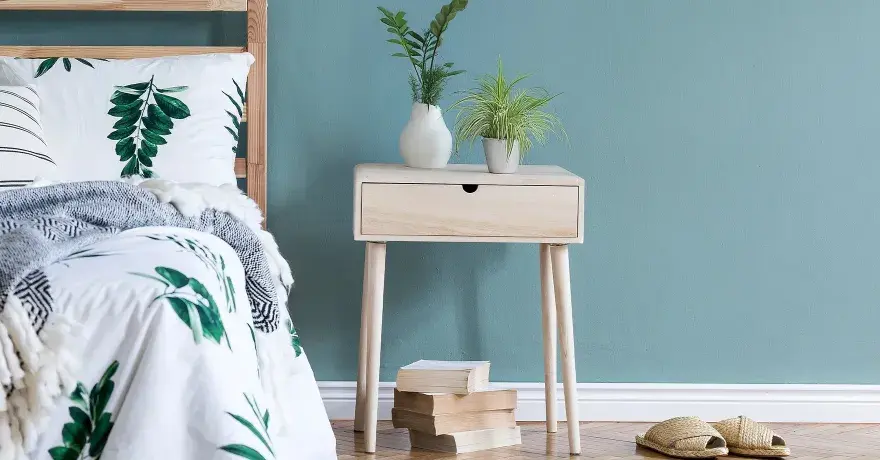 Ein Nachttisch DIY-Projekt aus hellem Holz mit einer Schublade, dekoriert mit Pflanzen und Büchern, neben einem Bett mit Blattmuster.