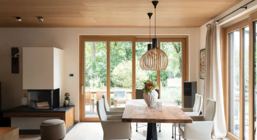 Ein Esszimmer mit Holzboden und Decke, wo sich eiche und buche kombinieren. Ein großer Holztisch steht vor bodentiefen Fenstern.