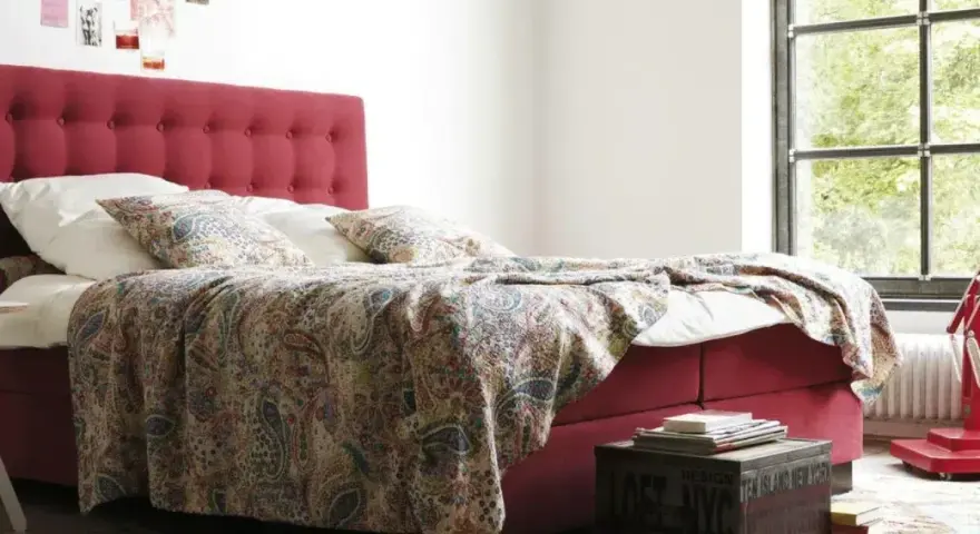 Rotes Boxspringbett mit gemustertem Überwurf und Kissen, ideal für Allergiker. Ein Nachttisch mit Büchern steht daneben.