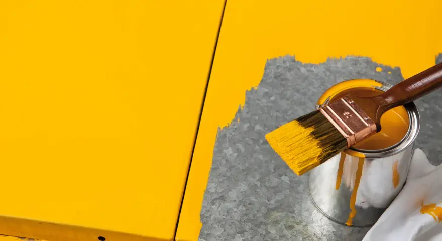 Gelbe Farbe tropft von einem Pinsel, der über eine Dose mit leuchtend gelbem Lack gehalten wird. Ein Teil einer gelben Oberfläche ist sichtbar, während daneben eine graue Fläche mit Farbe bedeckt wird.