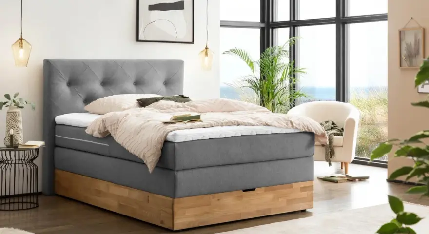 Gemütliches Boxspringbett ohne Lattenrost mit grauem Stoffbezug und Holzsockel. Einladend mit Decken und Kissen dekoriert, bereit für erholsame Nächte.