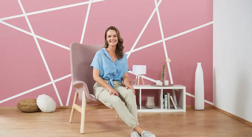 Frau sitzt vor einer rosa Wand, die mit weißen Linien diagonal gestrichen ist.