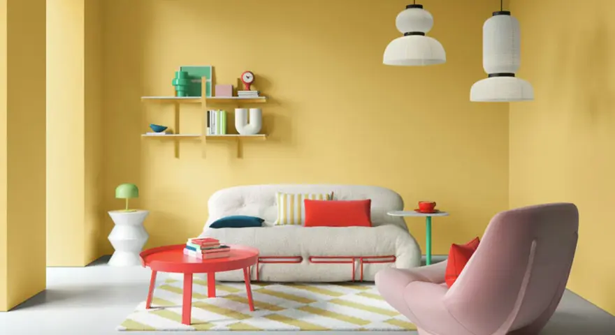 Ein Wohnzimmer mit gelben Wänden und einer auffälligen farb kombination aus Rot, Rosa und Weiß. Ein flauschiges Sofa und ein Sessel laden zum Entspannen ein.