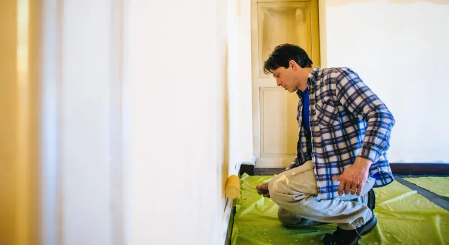 Mann kniet auf grüner Folie und trägt mit einer Rolle eine neue Farbe auf, um alte Latexfarbe zu entfernen und die Wand zu renovieren.