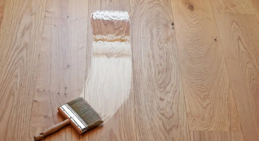 Breite Pinsel mit Holzgriff trägt Lack auf Parkett auf. Der Prozess, das Parkett zu versiegeln, zeigt den Unterschied zwischen unbehandeltem und frisch lackiertem Holz.