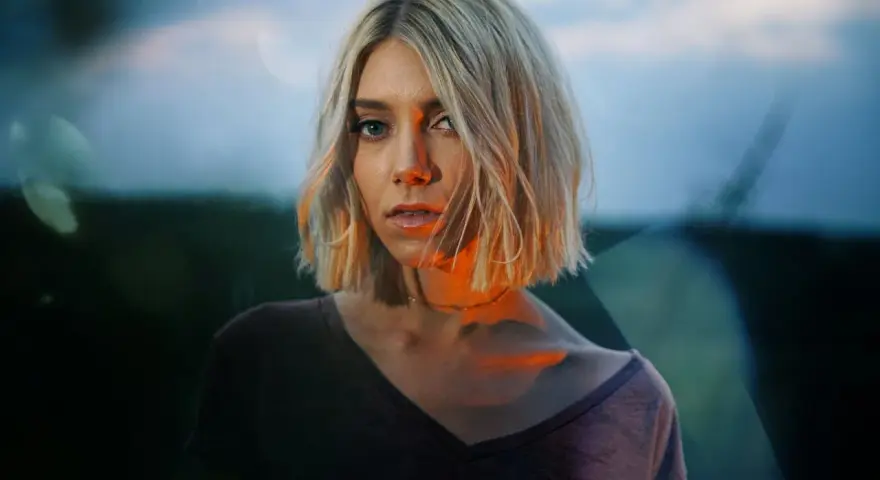 Blonde Frau mit Bob-Schnitt, deren Gesicht von warmem Licht beleuchtet wird, das sich mit dem blauen Abendhimmel mischt.