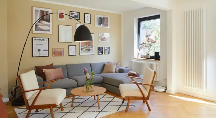 Gemütliches Wohnzimmer mit grauem Sofa, zwei Sesseln und vielen Bildern an der gelben Wand. Perfekt, um die Wand abzukleben und neu zu gestalten.