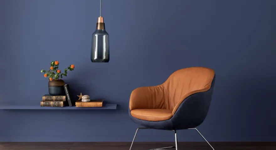 Ein Sessel mit braunem Leder und dunkelblauem Stoff vor einer blauen Wand. Orange Blumen auf einem Regal zeigen, welche Farbe passt zu Blau.