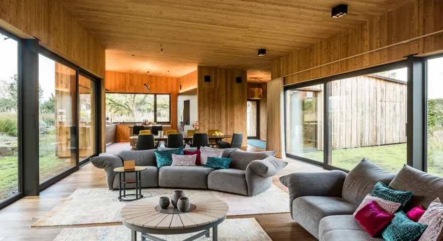 Gemütliches Wohnzimmer mit holzverkleideten Wänden und Decke, das zeigt, wie man eine holzdecke modern gestalten kann. Große Fensterfronten bieten Blick ins Grüne.