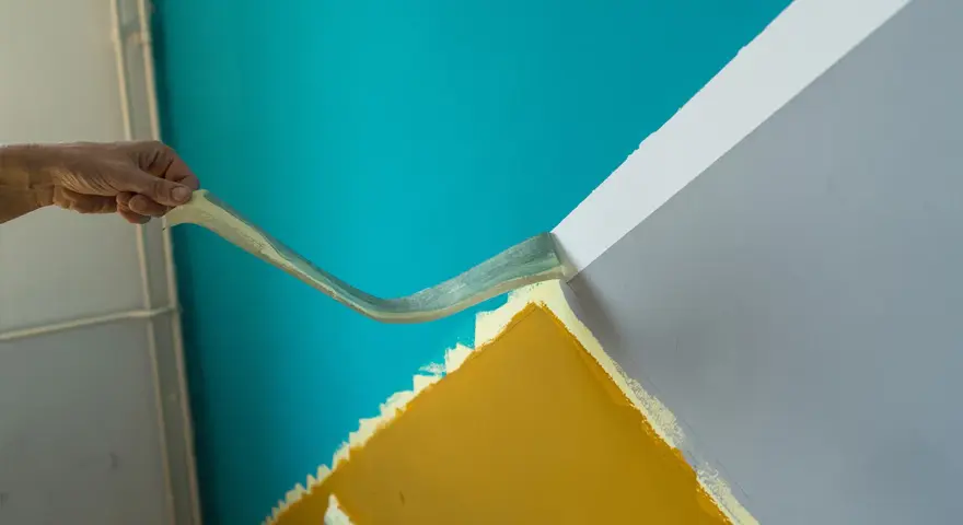Hand zieht Klebeband ab, um eine scharfe Kante beim Wand malen zu schaffen. Türkis trifft auf Grau und Ockergelb.