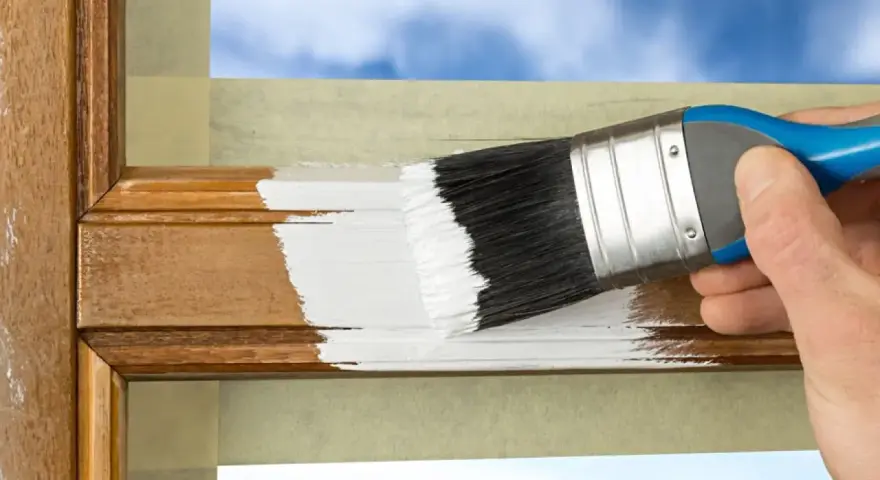 Hand streicht mit weißer Farbe über einen hölzernen Rahmen, um ihn aufzufrischen. Der Spiegelrahmen wird sorgfältig mit Klebeband geschützt.