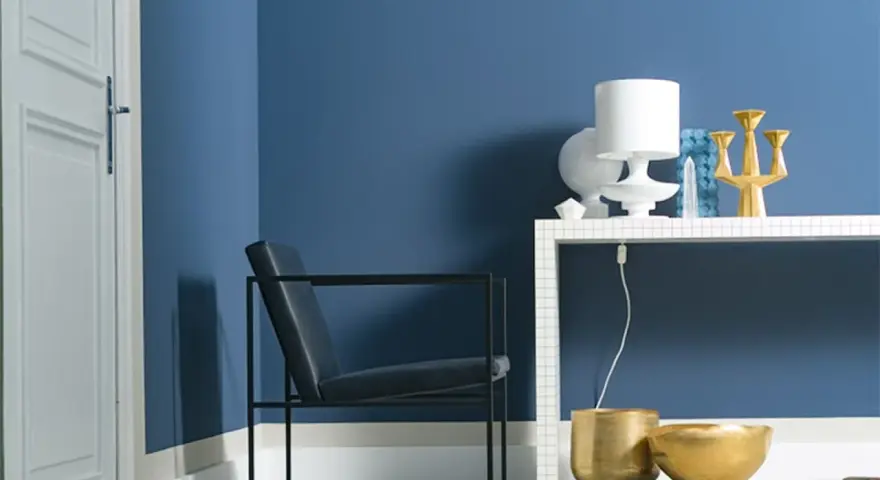 Ein Sessel vor einer Wand in moderner Wandfarbe. Auf einem Regal stehen eine Lampe, eine Vase und ein goldener Kerzenständer.