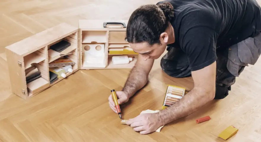 Mann repariert den Holzboden mit einem Reparaturstift und einer Farbkarte. Werkzeugkasten mit Zubehör daneben.