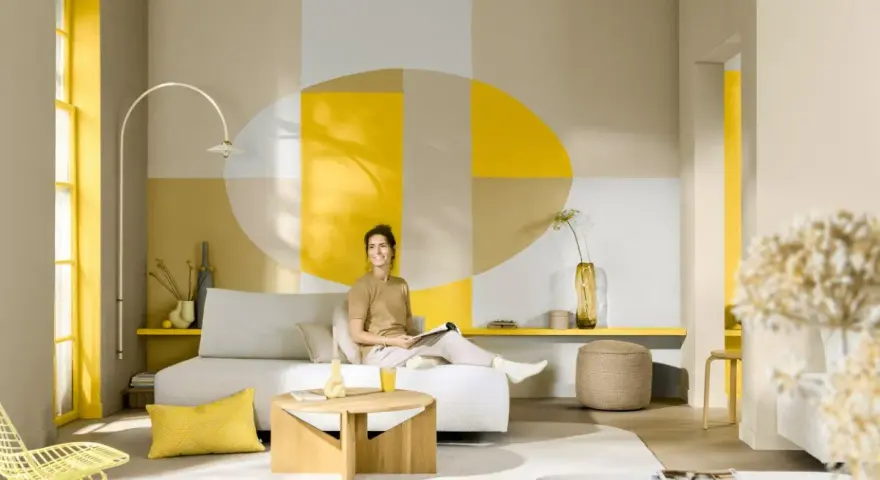 Eine Frau sitzt auf einem Sofa in einem hellen Wohnzimmer. Die Wand hinter ihr ist mit geometrischen Formen in Gelb, Beige und Grau gestaltet. Die Frage, welche Farbe passt zu Gelb, wird hier mit warmen Erdtönen und neutralen Farben beantwortet.
