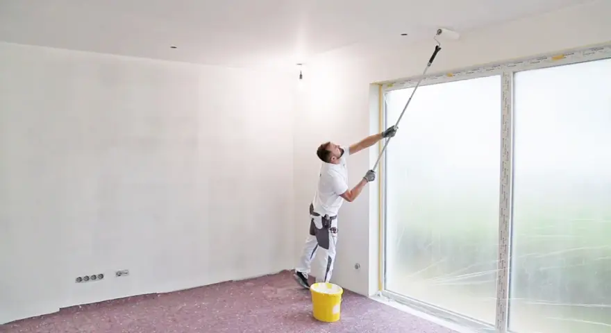 Maler streicht Decke und Wand mit langem Teleskoproller, um das Streichen ohne Abkleben zu ermöglichen. Gelber Farbeimer steht bereit.