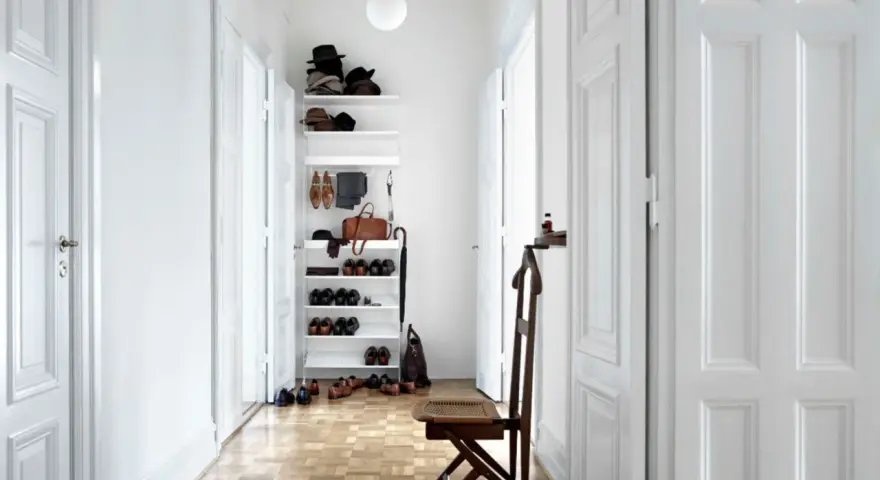 Schmaler Flur mit weißer Wandfarbe, Holzboden und einem Regal voller Schuhe und Hüte. Ein Stuhl steht bereit.