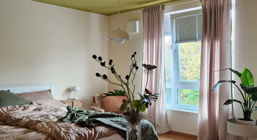 Gemütliches Schlafzimmer mit Bett in Khaki-Farbe, grüner Decke und rosa Vorhängen. Ein Blumenstrauß steht auf einem Holztisch.