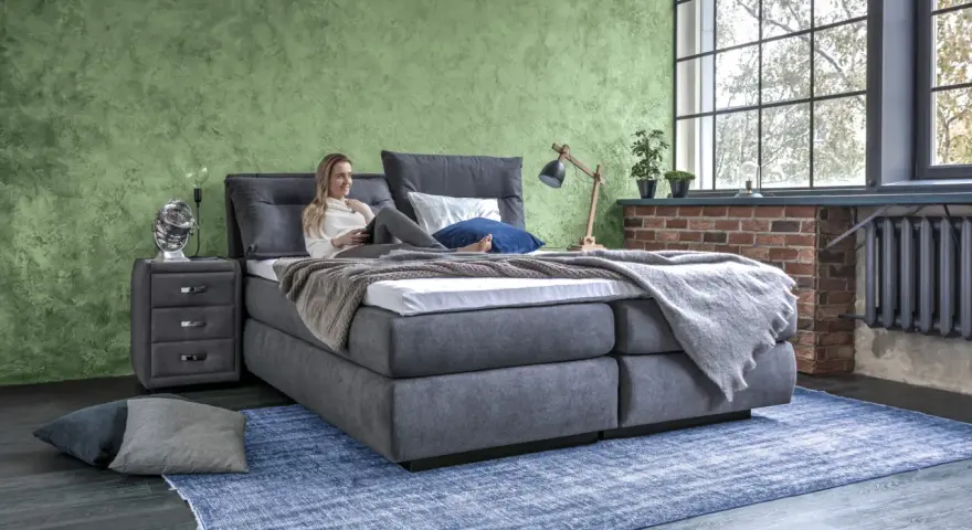 Frau entspannt auf einem Boxspringbett mit grauem Stoffbezug. Das Bett bietet hohen Komfort, was ein Boxspringbett ausmacht.