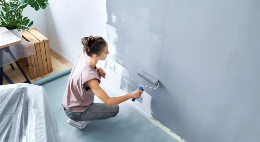 Frau streicht Wand grau. Nützliche **Wand streichen Tipps** für ein gleichmäßiges Ergebnis. Möbeltisch mit Folie abgedeckt.