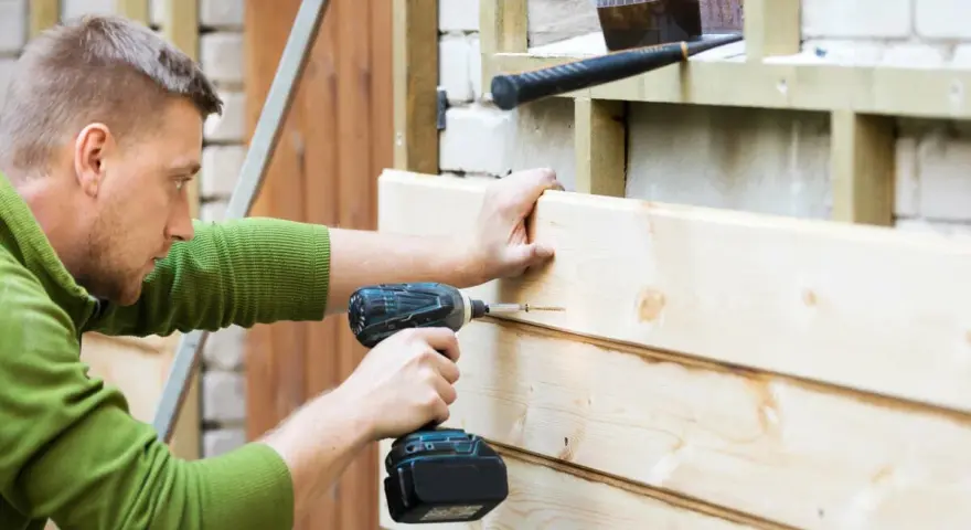 Mann renoviert Holzfassade, schraubt neue Bretter an. Werkzeug liegt bereit.