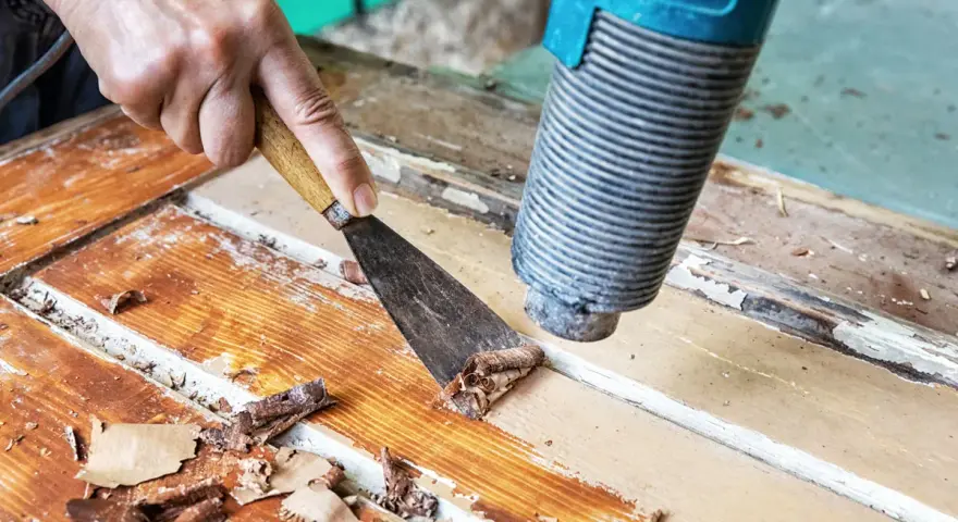 Farbe von Holz entfernen ohne Schleifen: Heißluftföhn und Spachtel lösen alte Lackschichten von einer Holztür.