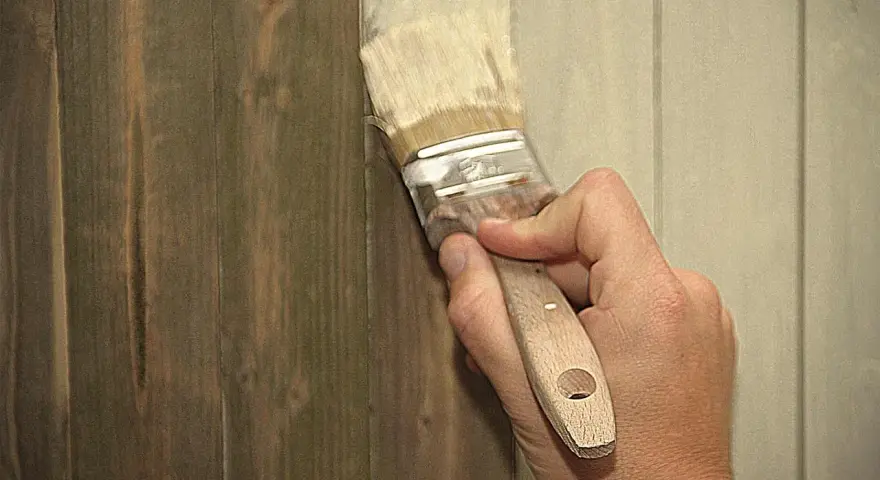 Hand streicht mit Pinsel über dunkles Holz, um es aufzuhellen. Ein Teil der Wand ist bereits heller gestrichen.