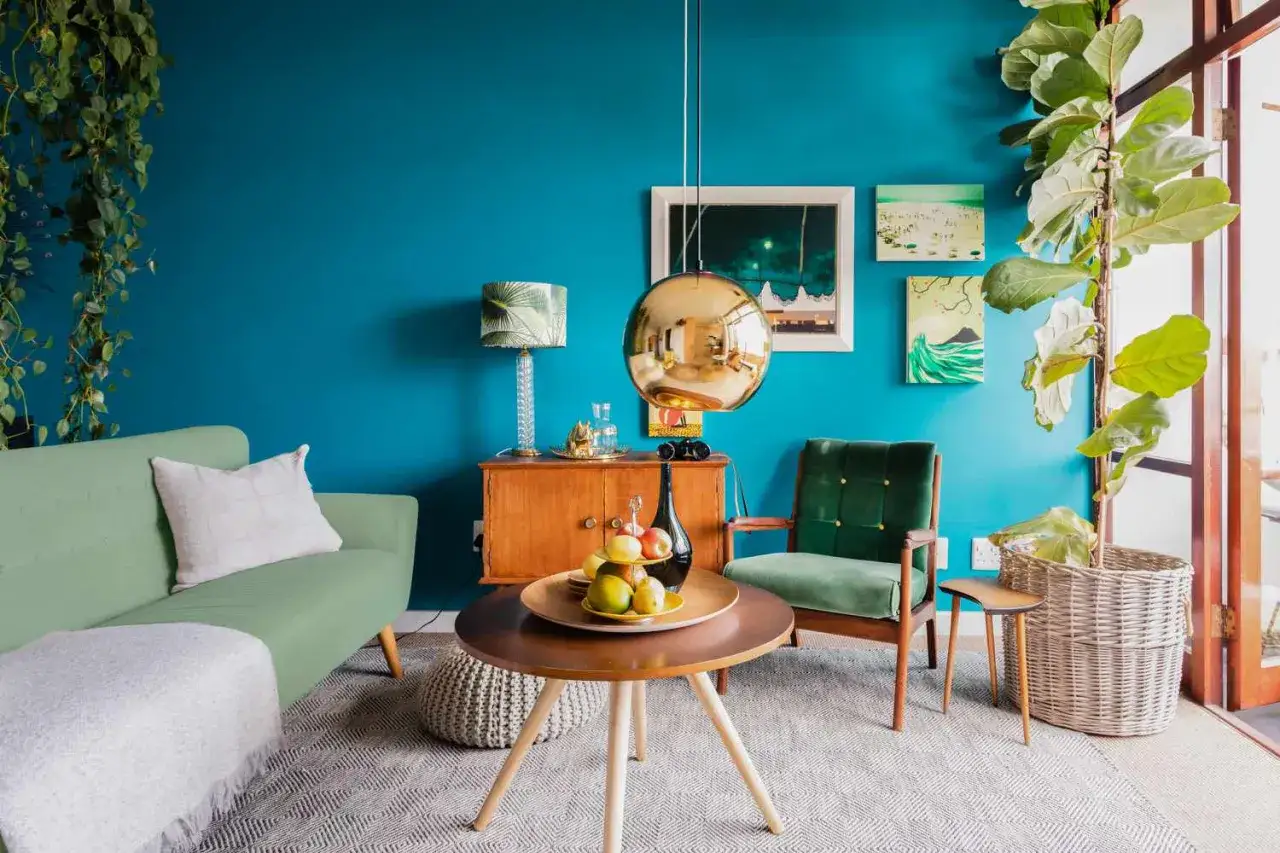 Ein lebendiges farbkonzept wohnzimmer mit türkisfarbener Wand, grüner Couch und Sessel. Goldene Lampe und Holzmöbel setzen Akzente.