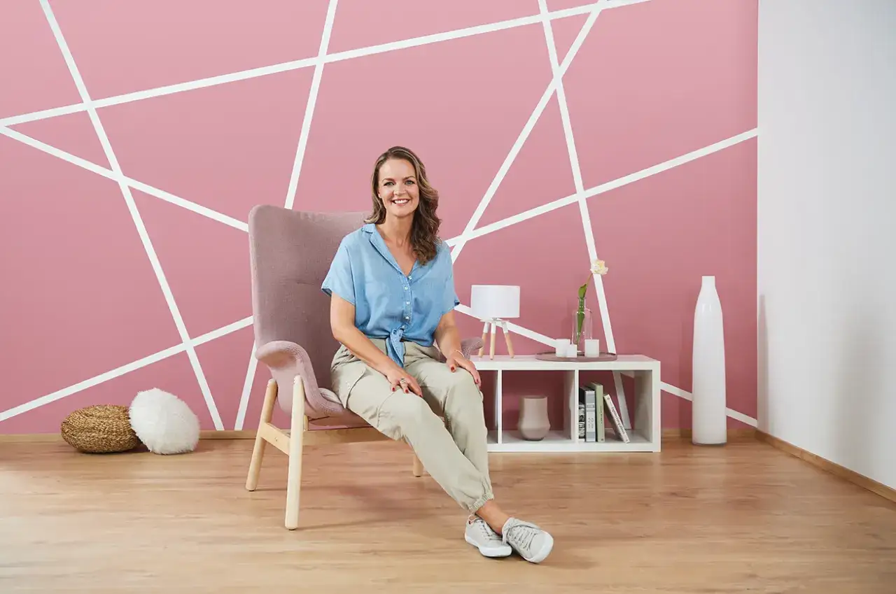 Frau sitzt vor einer rosa Wand mit geometrischer, kreativer Wandgestaltung. Ein Sessel und ein Regal mit Deko-Objekten vervollständigen das Bild.