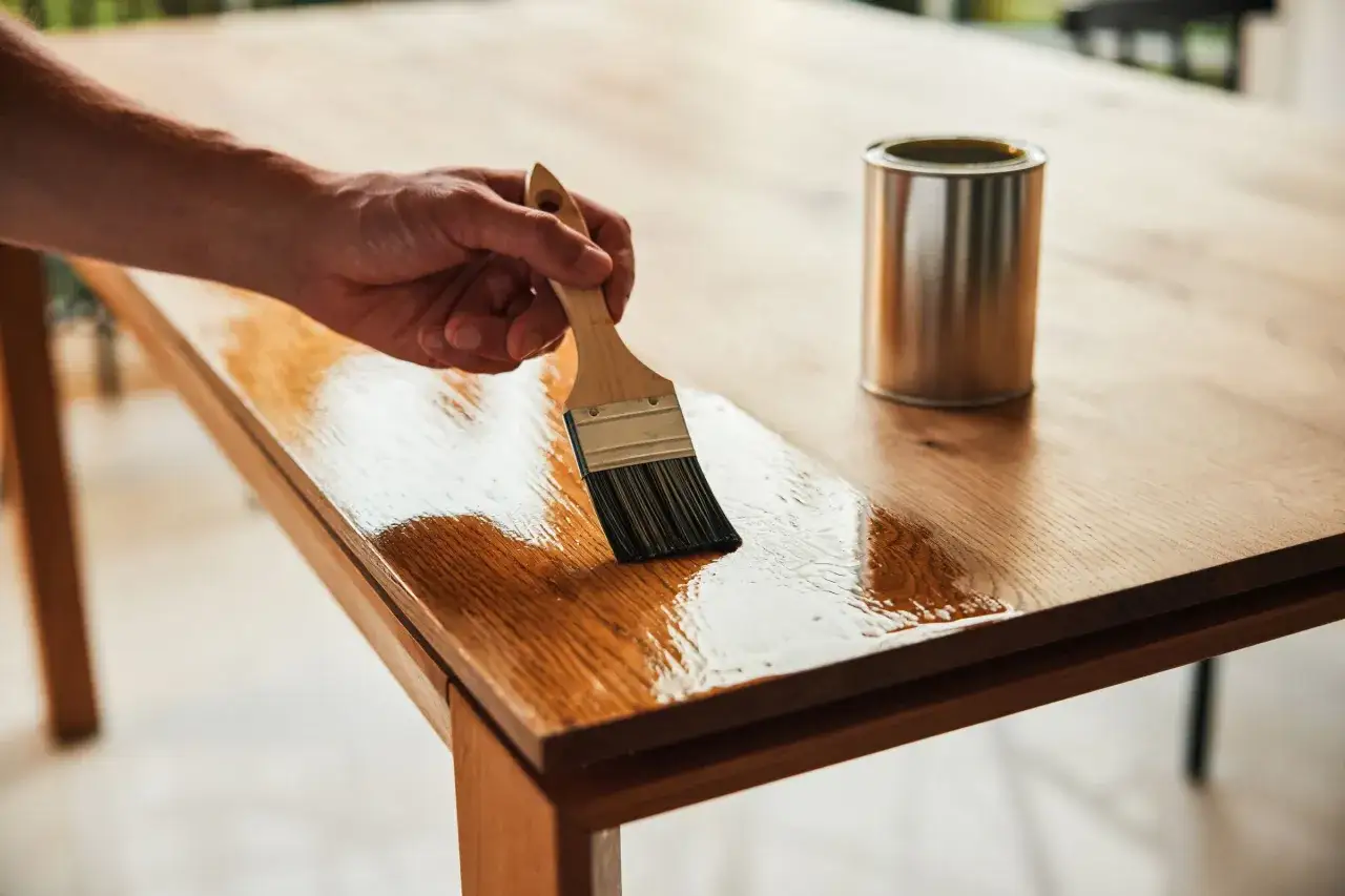 Hand streicht mit Pinsel über einen nassen Holztisch, Dose daneben. Pflege für den Holztisch.