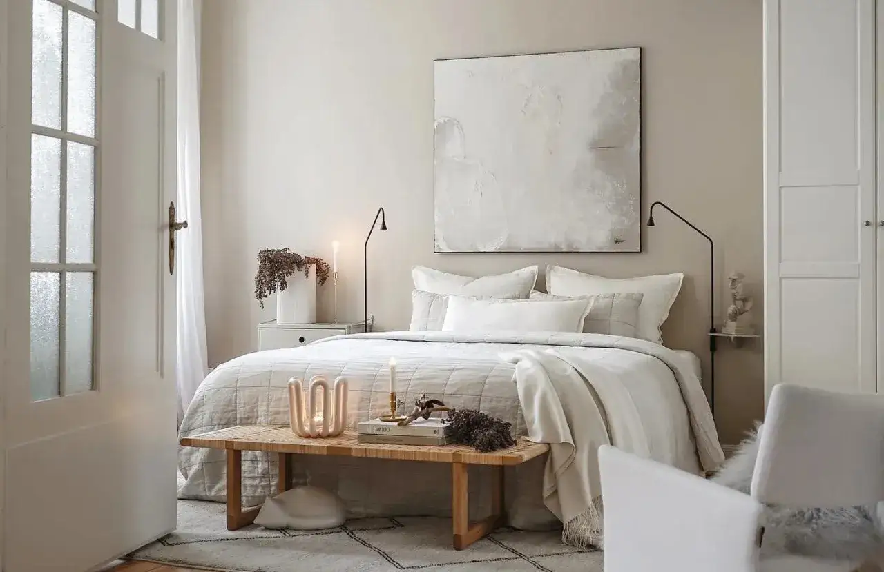Ein Schlafzimmer in sanften ecru farbe Tönen. Ein großes Bett mit vielen Kissen, ein Holzbettbank mit Kerzen und ein abstraktes Bild an der Wand.