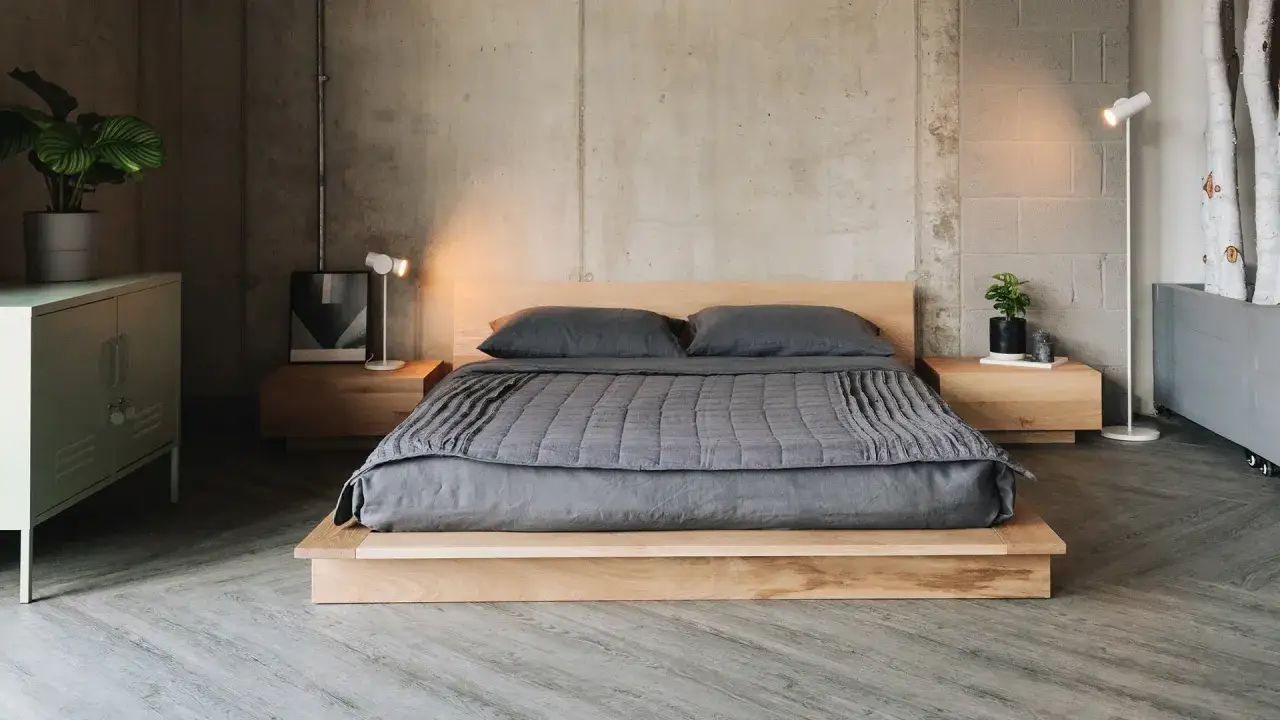 Ein Futonbett mit niedrigem Holzrahmen und grauer Bettwäsche steht vor einer Betonwand. Minimalistische Nachttische und Lampen ergänzen den modernen Look.