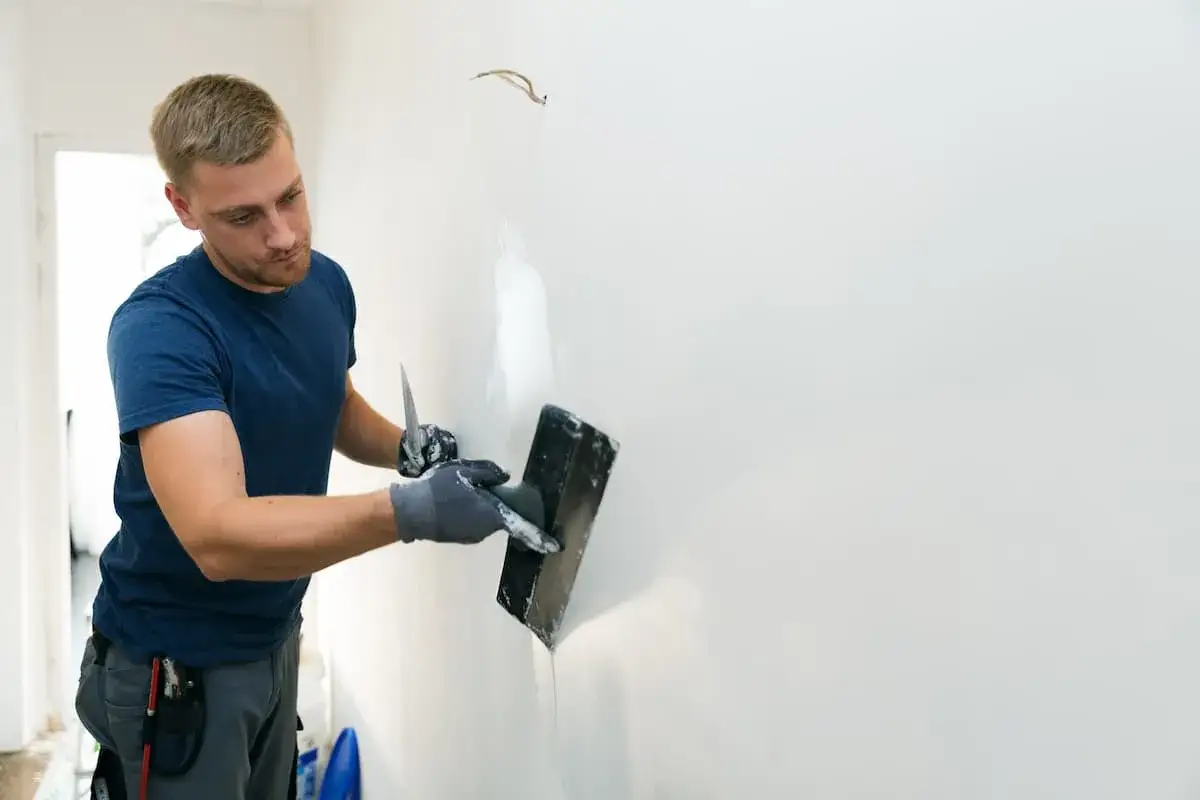 Mann mit Spachtel und Kelle bearbeitet eine Wand, um alte Latexfarbe zu entfernen und die Fläche für neue Farbe vorzubereiten.
