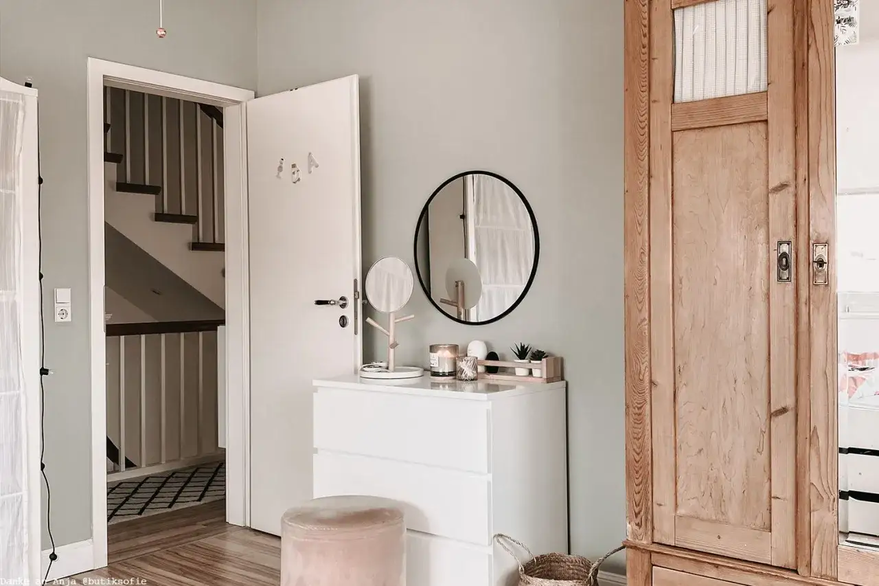 Ein zartes Altrosa passt wunderbar zu den hellen Holztönen und dem sanften Grau der Wand. Ein Hocker in Altrosa steht neben einer weißen Kommode.