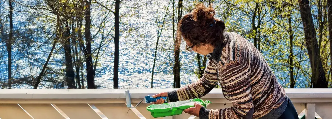 Frau streicht den Balkon weiß. Sie hält eine Farbrolle und eine grüne Farbwanne. Im Hintergrund glitzert Wasser zwischen Bäumen.