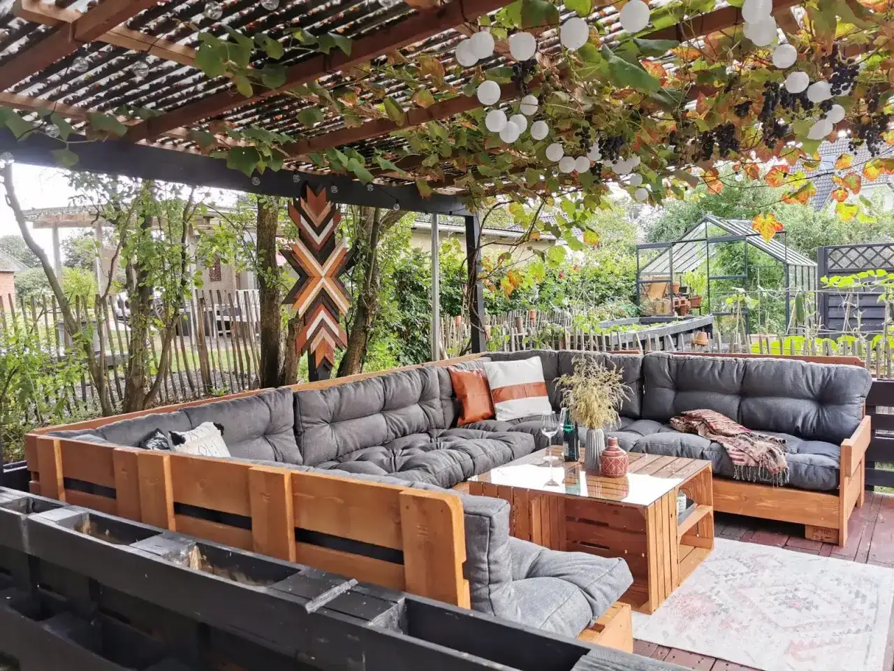 Gemütliche Paletten Lounge Ideen für den Garten: Eine Eckcouch aus Paletten mit grauen Polstern und Holzakzenten, überdacht von einer Pergola mit Weinreben und Lichterketten.
