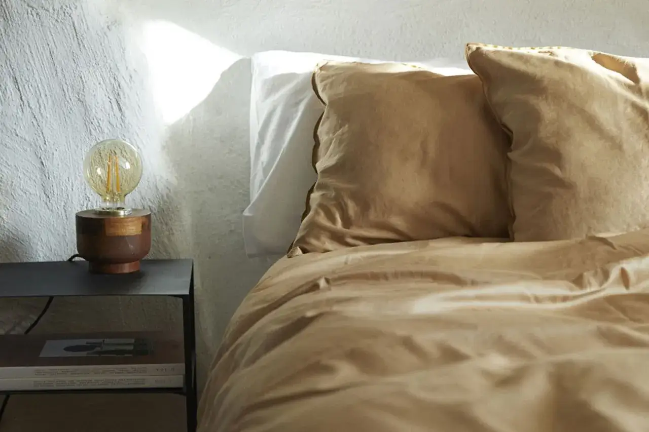 Ein Bett mit ockerfarbener Bettwäsche und Kissen, daneben ein Nachttisch mit Lampe und Büchern.