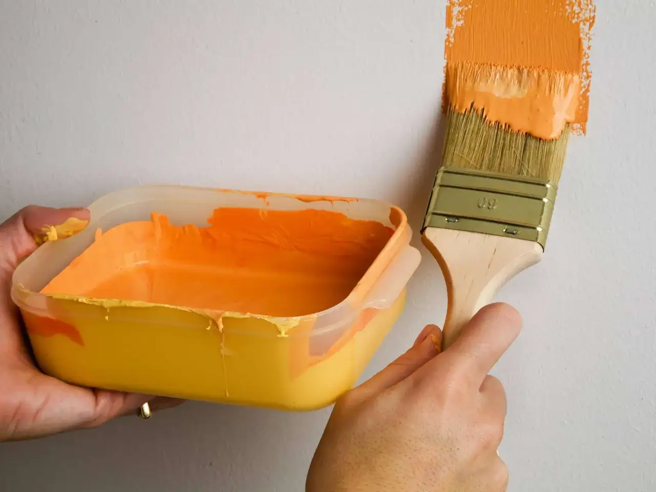 Orange Farbe wird mit einem Pinsel auf eine wei&szlig;e Wand gestrichen, ohne abzukleben. Ein Beh&auml;lter mit Farbe ist in der anderen Hand.