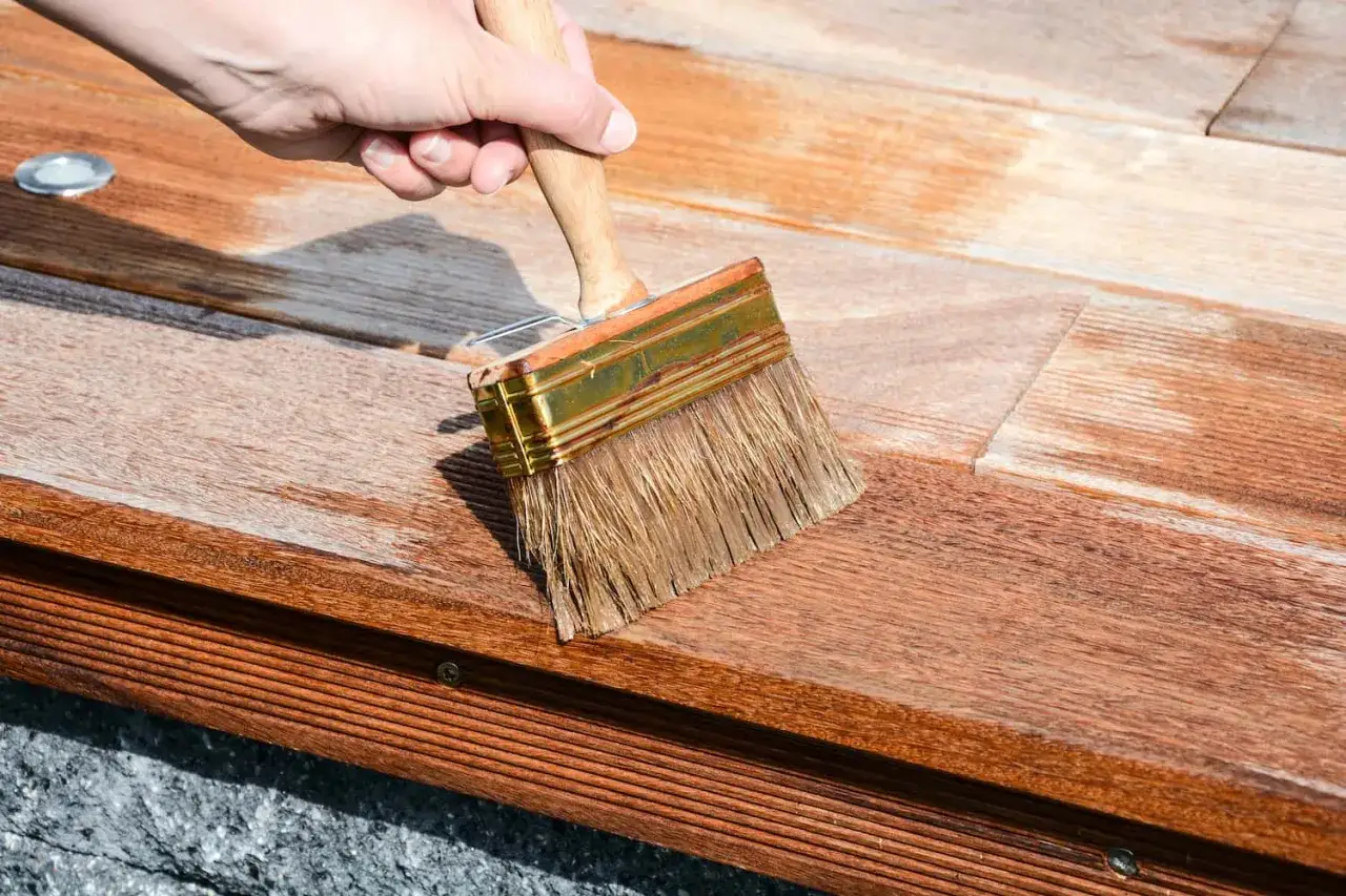 Hand streicht mit breitem Pinsel über ausgeblichenes Holz, um es aufzufrischen. Die Holzoberfläche wird sichtbar dunkler und erhält neuen Glanz.