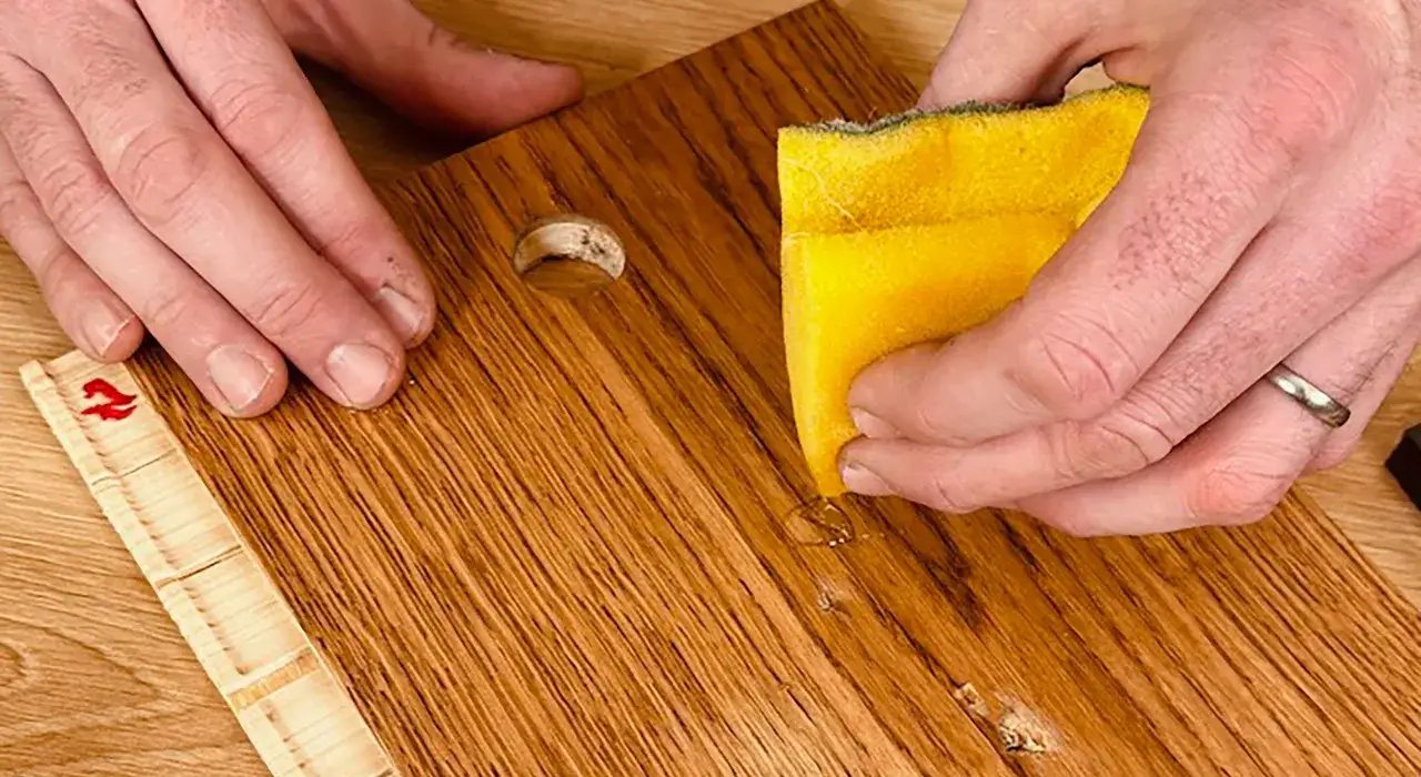 H&auml;nde reparieren einen Holzboden. Eine Hand h&auml;lt einen Schwamm, um eine Stelle zu reinigen, w&auml;hrend die andere Hand einen Holzd&uuml;bel positioniert.