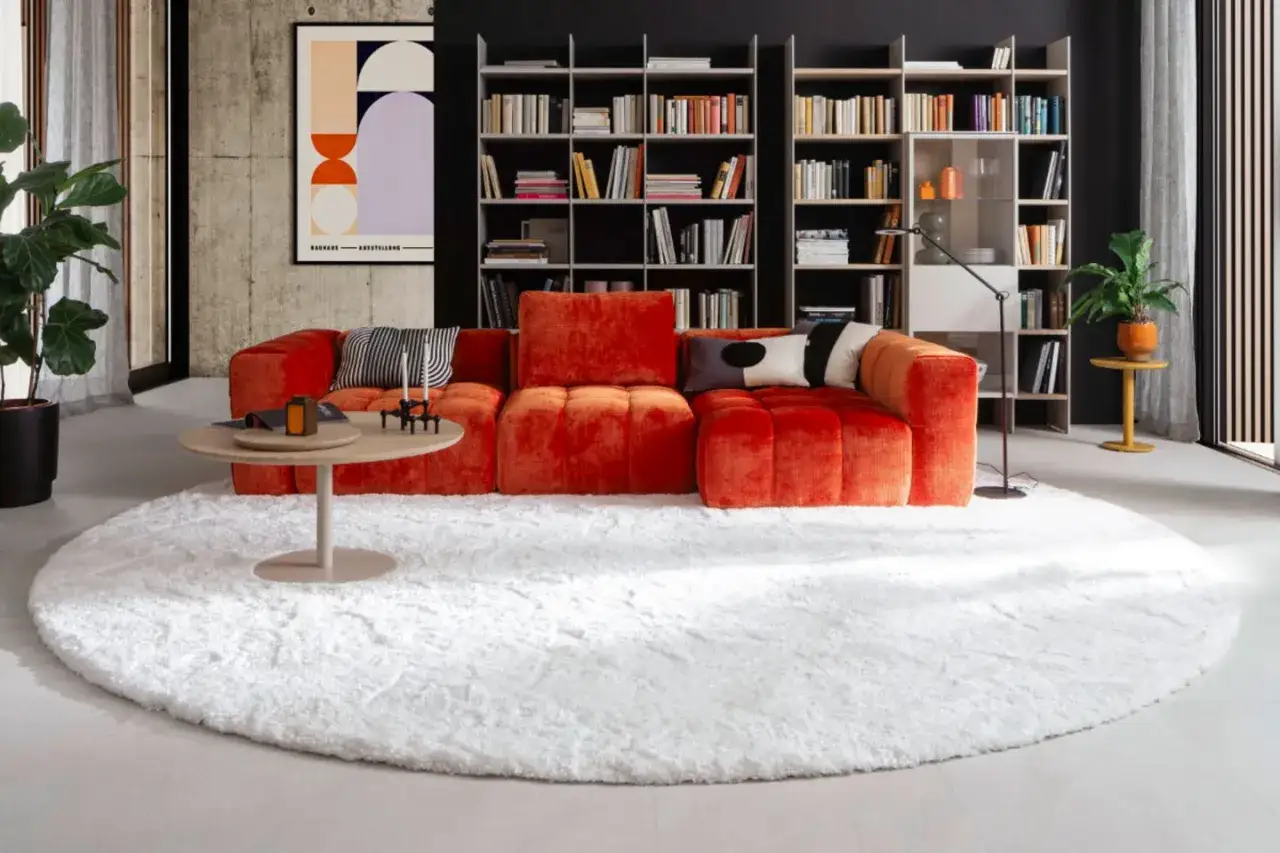 Ein flauschiger weißer Teppich bildet die Basis für ein modernes Wohnzimmer. Ein leuchtend orangerot Sofa ist darauf richtig platziert, mit einem runden Couchtisch davor.