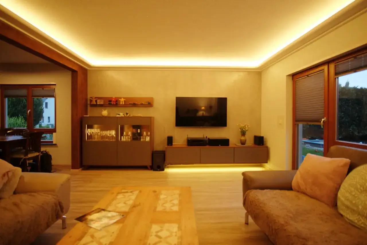 Ein Wohnzimmer mit moderner Beleuchtung, die eine holzdecke modern gestalten lässt. Gemütliche Sofas und ein TV-Schrank laden zum Entspannen ein.