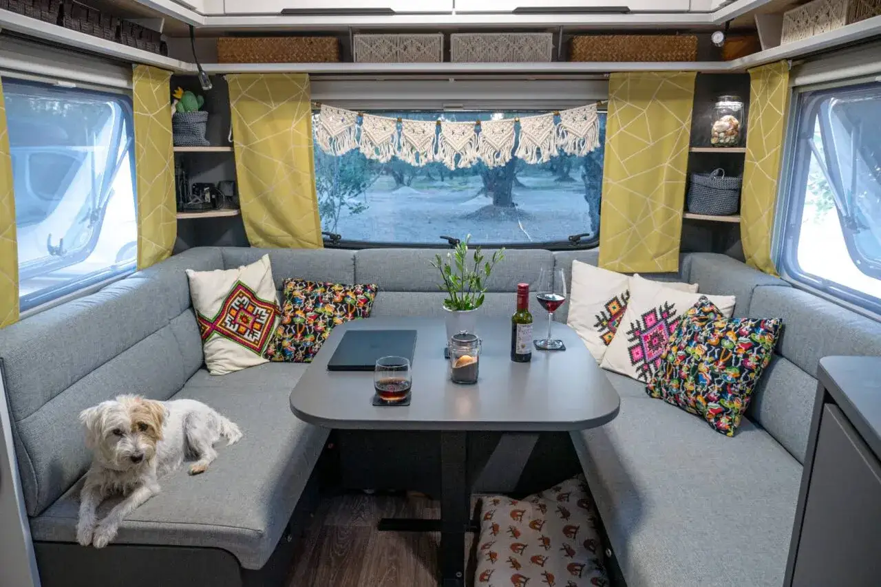 Gemütliche Wohnwagen-Inneneinrichtung mit U-förmiger Sitzbank, bunten Kissen, Tisch mit Wein und einem Hund. Tolle Ideen für Wohnwagen innen.