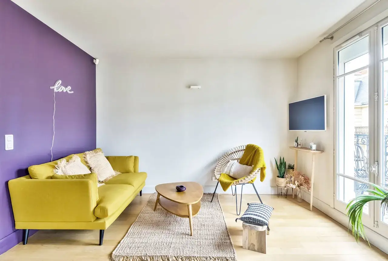 Ein gem&uuml;tliches Wohnzimmer mit einer lila Wand, einem gelben Sofa und einem Korbsessel mit gelber Decke. Ein kleiner Holztisch steht davor.