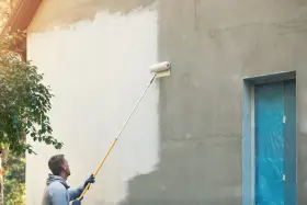 Mann streicht mit einer Teleskoprolle die Hausfassade weiß. Die rechte Seite ist noch grau, die linke bereits frisch gestrichen.