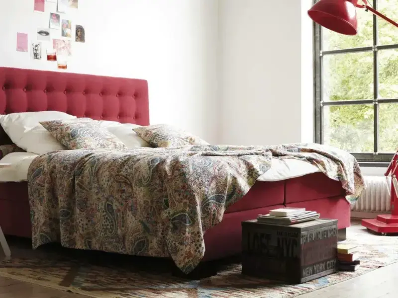 Rotes Boxspringbett mit gemustertem Überwurf und Kissen, ideal für Allergiker. Ein Nachttisch mit Büchern steht daneben.